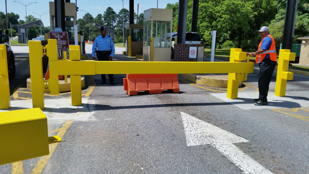 Yellow crash-rated vehicle barrier across facility entrance with a security guard standing nearby.