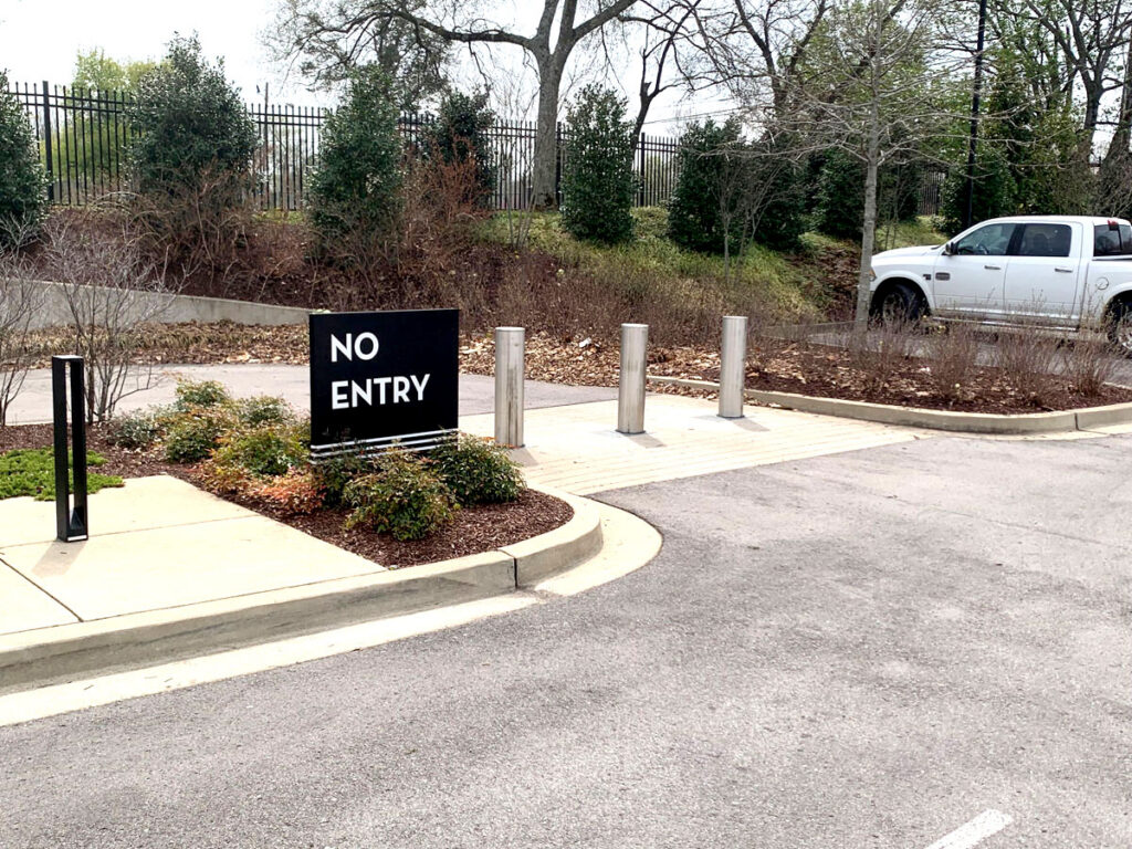 Three stainless steel fixed bollards blocking a paved drive with a “No Entry” sign.